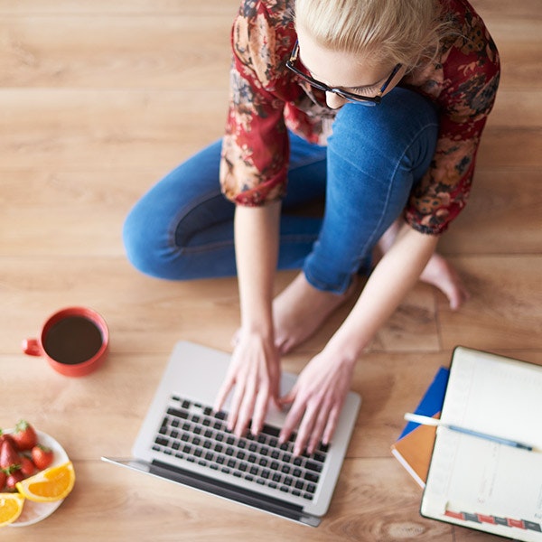 woman using her laptop while sitting on a hardwood floor, next to a cup of coffee, bowl of fruit, and pile of notebooks